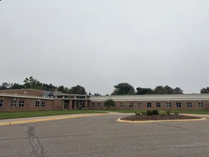 A one-story brick school building with many windows, situated behind a roundabout driveway and a landscaped area with mulch and small shrubs under a cloudy sky.