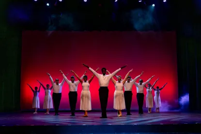A group of dancers in coordinated white outfits perform on stage, standing in rows with arms raised and a red-lit background behind them. The stage is illuminated with spotlights above.