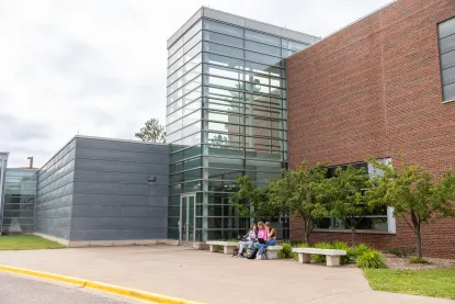 Three people sit on a bench outside a modern building with glass and brick walls. Trees and greenery are near the entrance, and the sky is overcast.