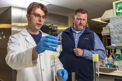 A student in a lab coat and faculty member investigating beakers filled with liquid.