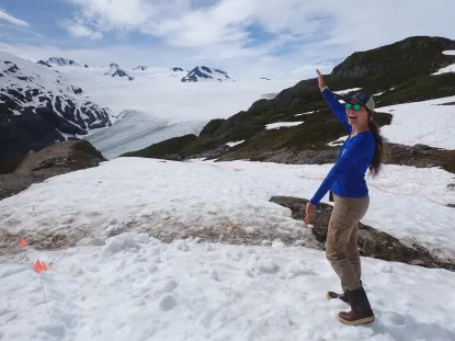 A student stands on the top of a mountain, gesturing out the a bigger mountain in the distance.