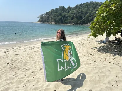 NMU student stands on a beach holding the NMU flag.