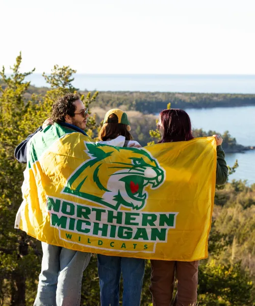 Three students on top of a mountain holding a Northern Michigan University Wildcat flag
