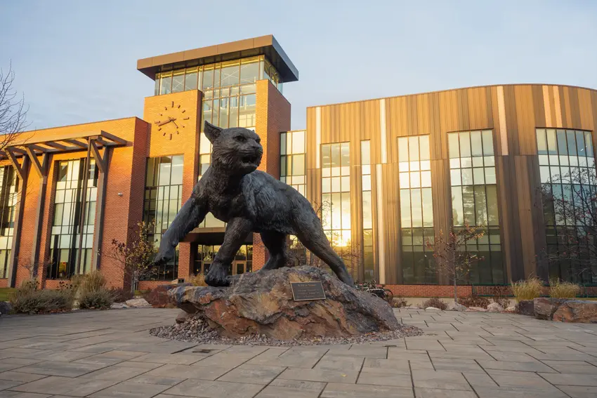 The NMU wildcat statue outside of Jamrich Hall with the sun rising on the large windows of the hall.