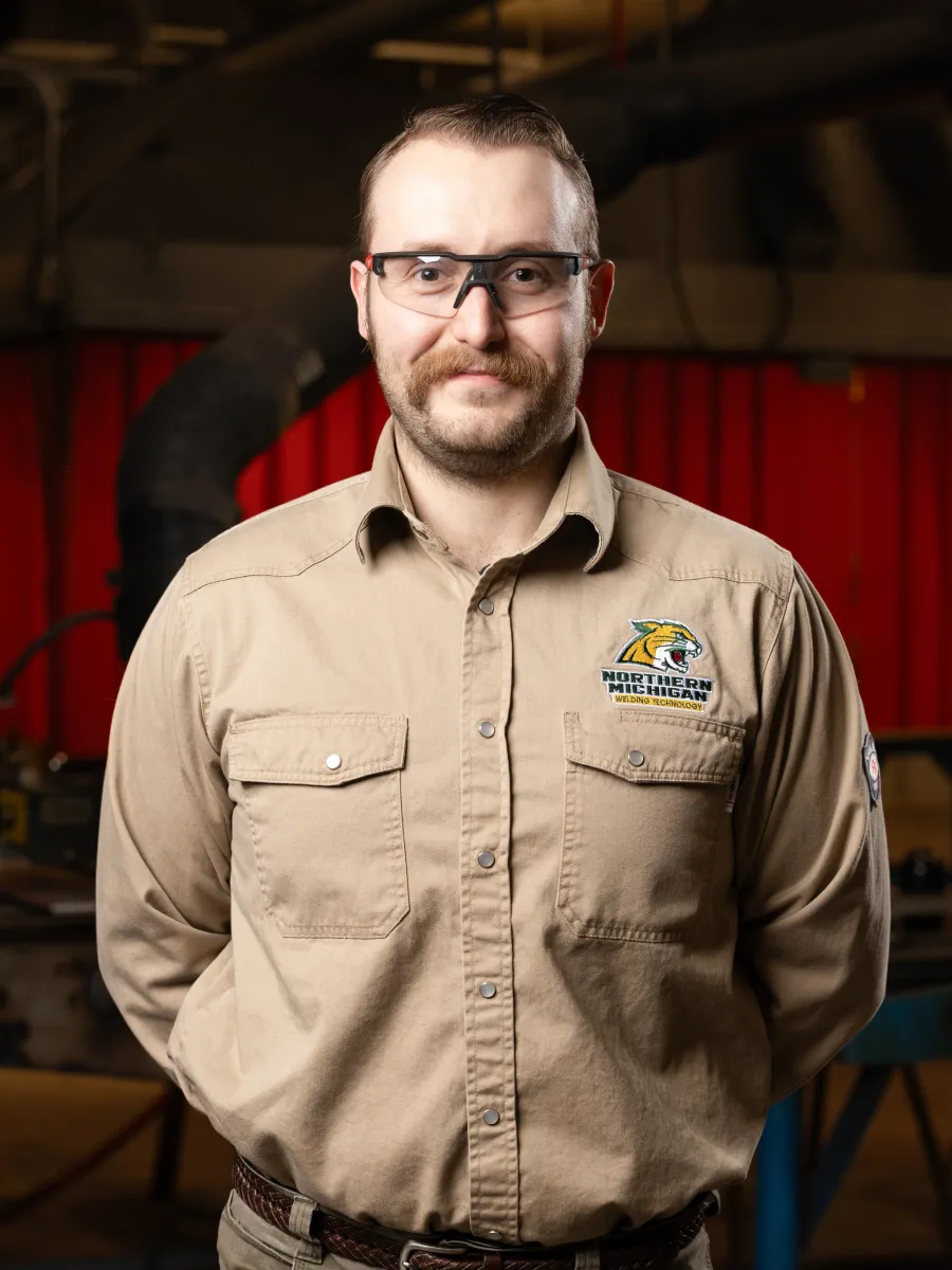 Male with facial hair wearing safety glasses and welding burn shirt with NMU Welding logo smiles at camera
