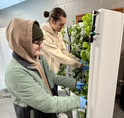 One male student and one female student work with the produce growing in upright hydroponic indoor farming stations.