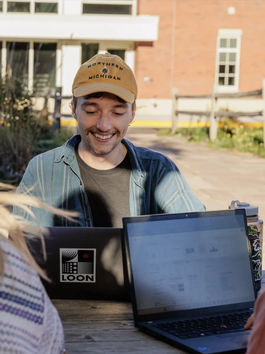 Male wearing a Northern Michigan University hat sits outside on his lap top