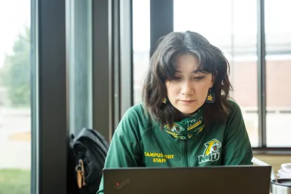 Female student in green NMU jacket sits at table with laptop in front of her