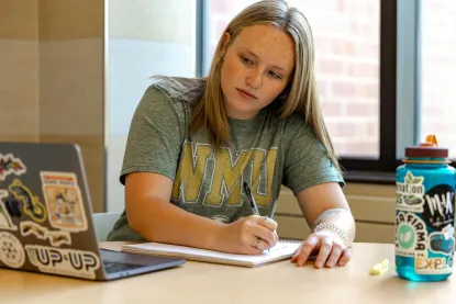 Student writing in a notebook while looking at a laptop.