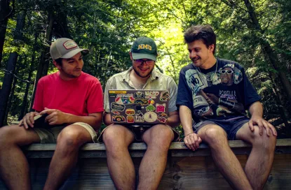 Three males sit on a wooden bridge in the forest with a laptop in the middle of them