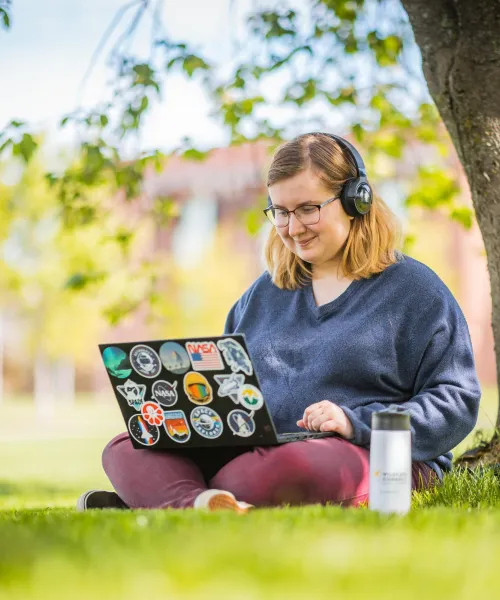 Student sitting under a tree on NMU’s campus, working on a laptop.