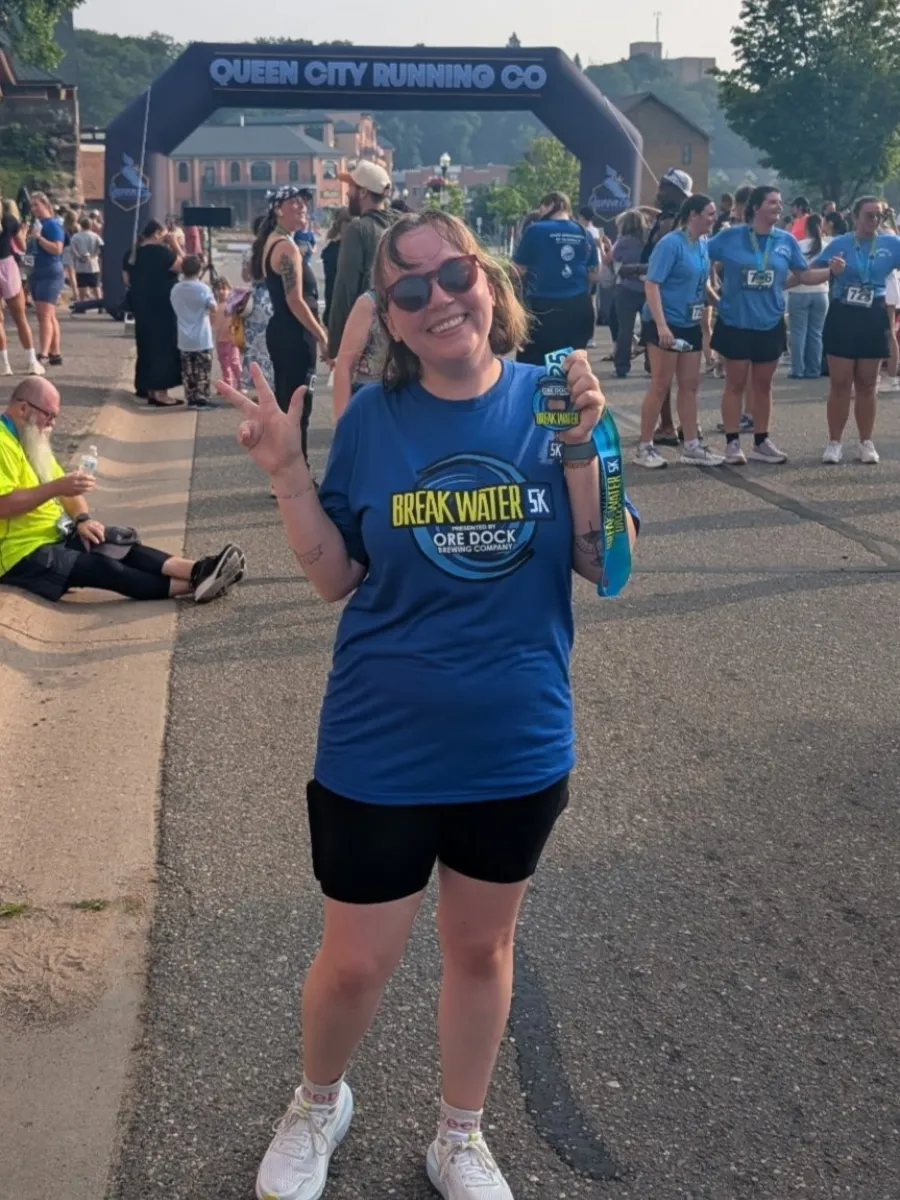 Photo of woman giving thumbs up and holding a medal by a finish line, wearing a blue shirt and black running shorts.