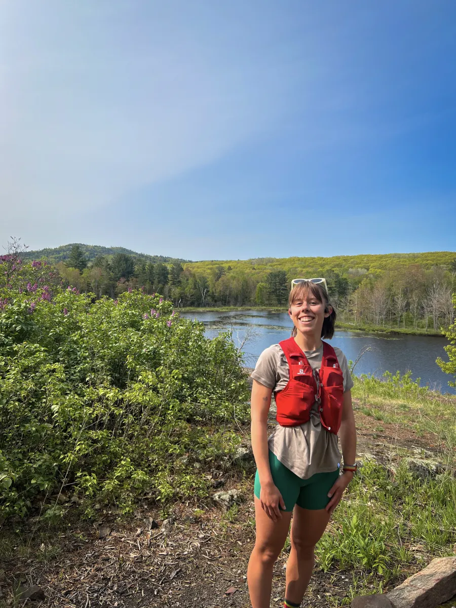 Woman in orange hydration vest and running attire standing in scenic area with mountain in background.