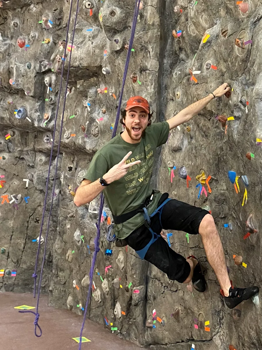 Image of man on climbing wall, giving peace sign with one hand.