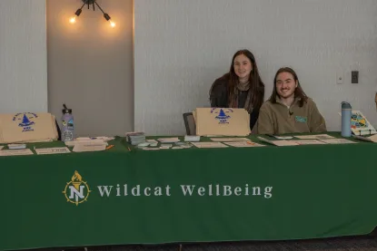 Two students behind table that has green Wildcat WellBeing tablecloth on it as well as tote bags and resouces.