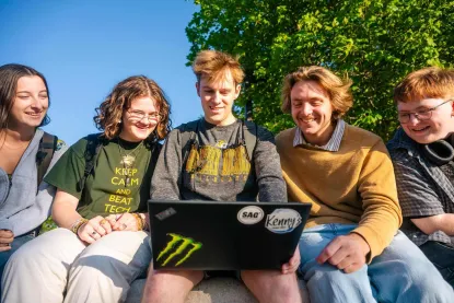 Five students sitting on a bench looking at the center student typing on a laptop.